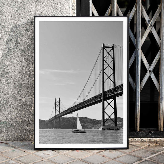 Lisbon Bridge Black and White Poster by Raphael Alves featuring a framed photograph of a suspension bridge and sailboat in the water, framed and displayed in home decor