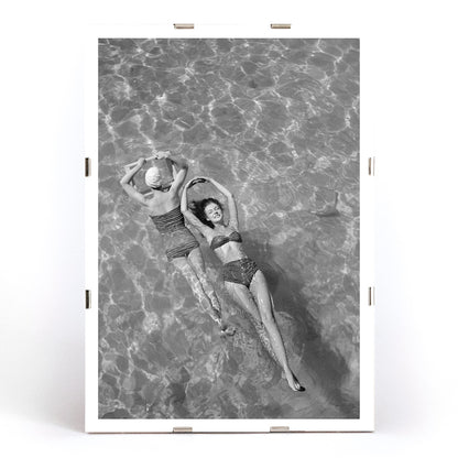 Fashion models in vintage bathing suits floating in a swimming pool artwork by Toni Frissell, framed and displayed in modern living room decor
